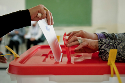 Hand putting voting card into ballot box, other hands hold open ballot box