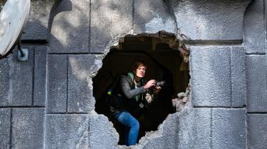 A photographer in a war zone looking through a hole blasted in a wall