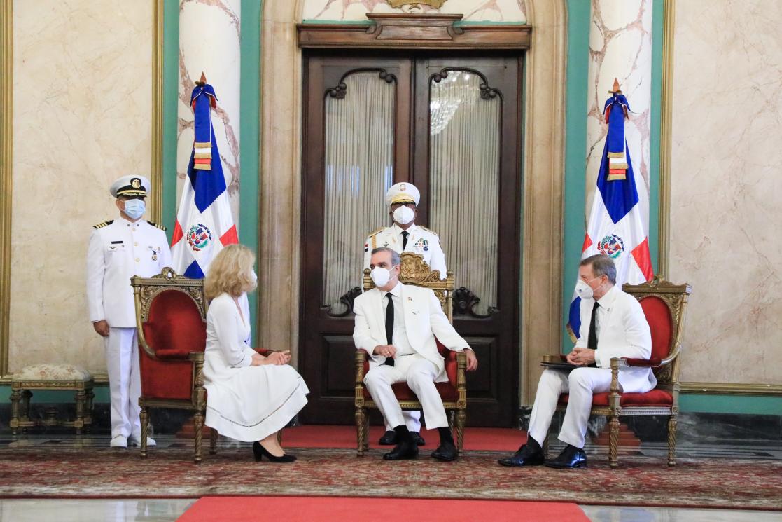 Three people dressed in white talking sitting on three armchairs, guarded by two military personnel
