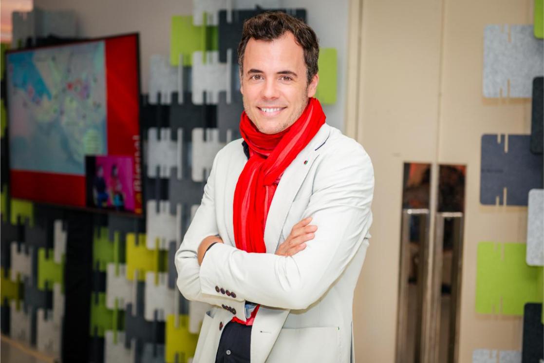 A man in a red scarf and white jacket, smiles and poses.