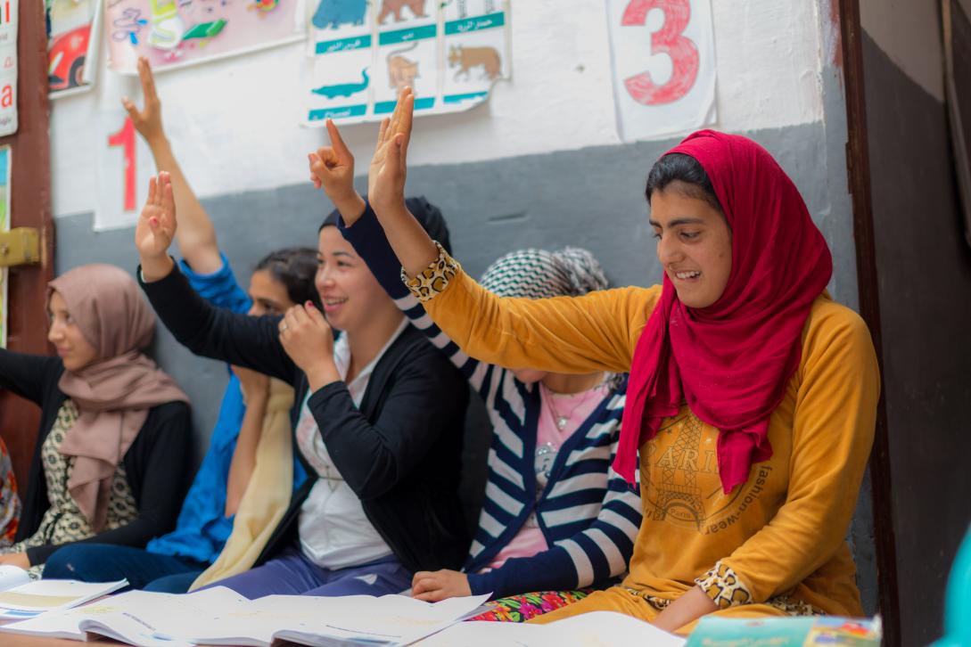 cinq jeunes filles avec les mains levées dans une salle de classe avec derrière un mur avec des dessins scolaires