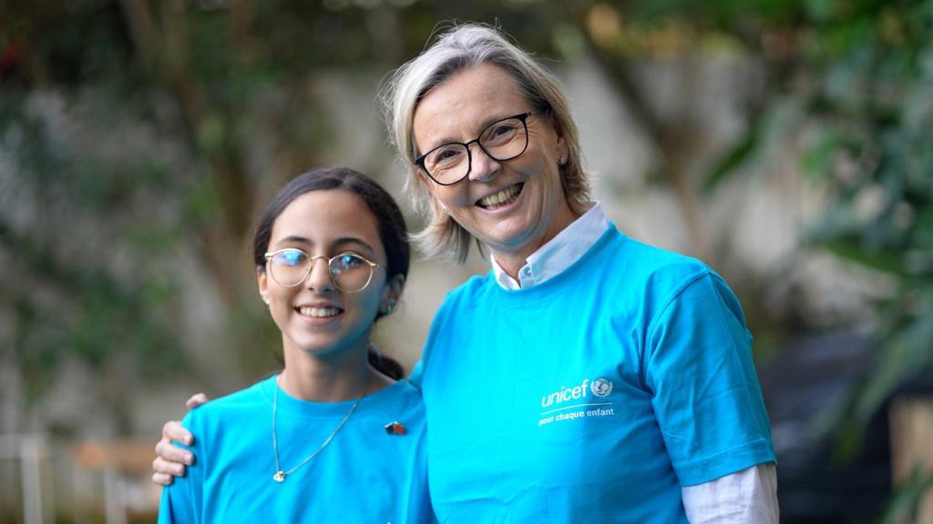 A gauche une enfant avec des lunettes habillée avec un T-shirt bleu et à droite une femme habillée avecc un T-shirt bleu qui a le logo de l'Unicef